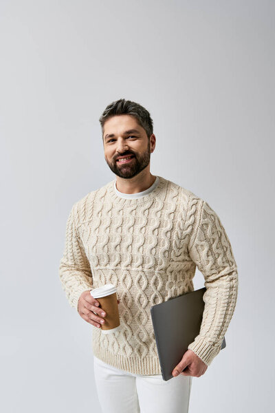 A bearded man in a white sweater holds a coffee cup and a laptop against a grey background.