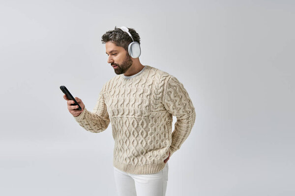 A bearded man in a white sweater, wearing headphones, engrossed in his cell phone screen against a grey backdrop.