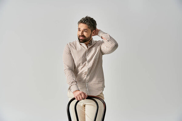 A stylish man with a beard sitting confidently on top of a chair against a grey background in a studio setting.