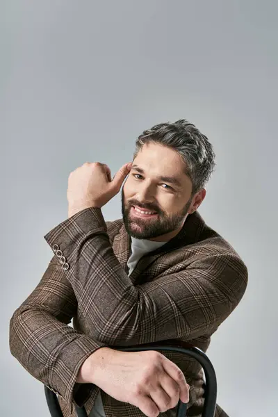 stock image A stylish man with a beard sits in a chair, hand on head, lost in thought, exuding elegance and sophistication.