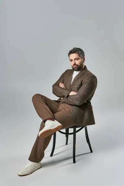 stock image A bearded man sits with arms crossed, exuding strength and poise in elegant attire against a grey studio backdrop.