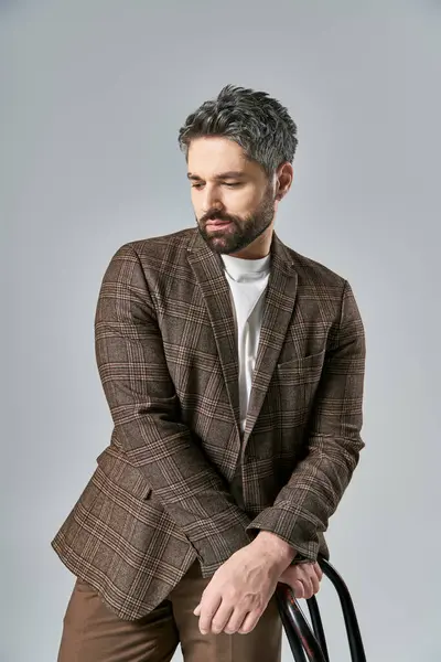 Stock image A bearded man in a brown jacket and pants poses with a black chair in a studio setting against a grey background.