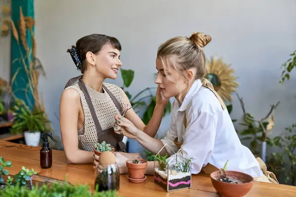 İki sanatkar kadın saksı bitkileriyle çevrili bir masada oturur ve sevgi dolu bir birliktelik yaratırlar..