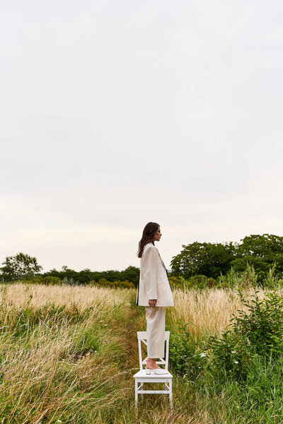 A beautiful young woman in white attire stands on a chair, enjoying the summer breeze in a field.