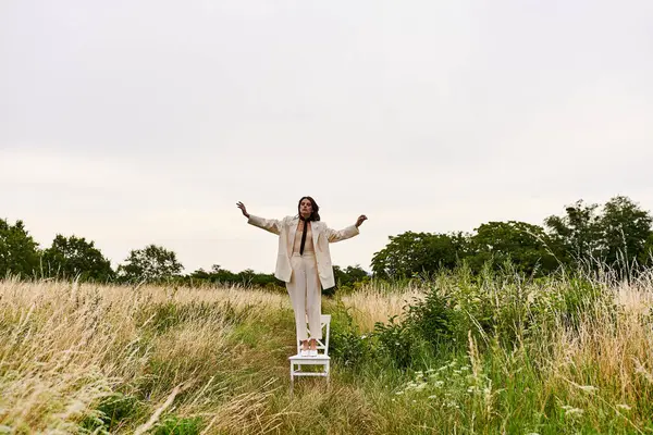 stock image A beautiful young woman in white attire stands confidently on a chair in a lush field, enjoying the summer breeze.