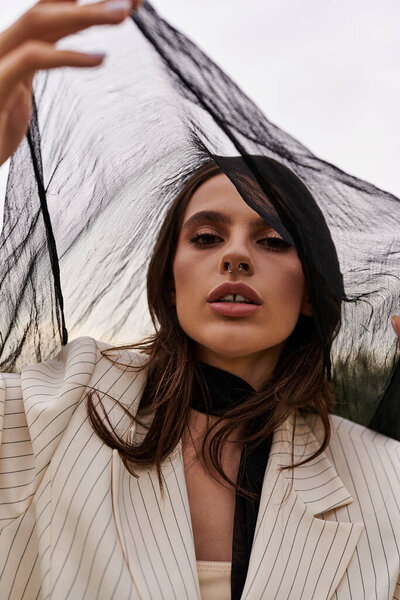 A stylish young woman in a suit stands in a field, holding an umbrella as she enjoys the summer breeze.