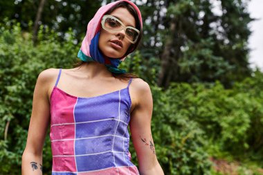 Parlak elbiseli ve güneş gözlüklü güzel bir genç kadın, doğada yaz esintisinin tadını çıkarıyor..