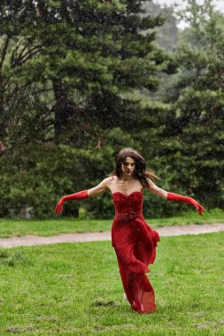 Çarpıcı kırmızı elbiseli ve uzun eldivenli genç bir kadın yağmurda zarif bir şekilde koşuyor ve doğal elementleri kucaklıyor..