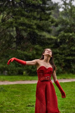 Kırmızı elbiseli ve uzun eldivenli çekici genç bir kadın yağmurda zarif bir şekilde duruyor ve yaz yağmurunun tadını çıkarıyor..