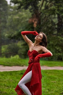 Kırmızı elbiseli ve uzun eldivenli genç bir kadın yağmurda zarif bir şekilde duruyor, doğadaki yaz esintisini kucaklıyor..
