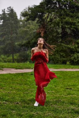 Kırmızı elbiseli ve uzun eldivenli büyüleyici genç bir kadın zarif bir şekilde yağmurda duruyor, yaz duşunu kucaklıyor..