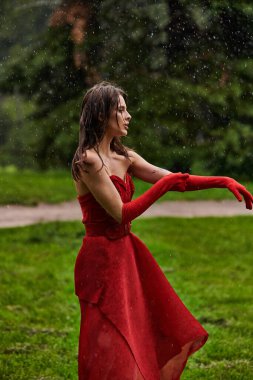 Dalgalı kırmızı elbiseli ve uzun eldivenli genç bir kadın yaz yağmurunda zarif bir şekilde dans ediyor. Duruş ve zarafeti simgeliyor..