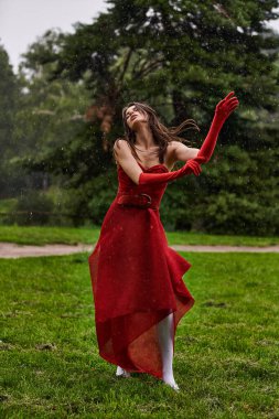 Kırmızı elbiseli genç bir kadın yağmurda zarif bir şekilde duruyor, doğal bir ortamda yaz melteminin tadını çıkarıyor..