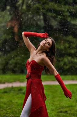 Kırmızı elbiseli ve uzun eldivenli çekici genç bir kadın yağmurda neşeyle dans ediyor, yaz esintisini kucaklıyor..