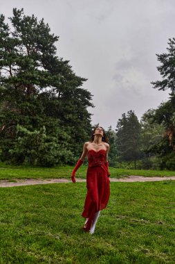 Canlı kırmızı elbiseli ve uzun eldivenli zarif genç bir kadın yemyeşil çimlerin arasında duruyor, yaz melteminin tadını çıkarıyor..