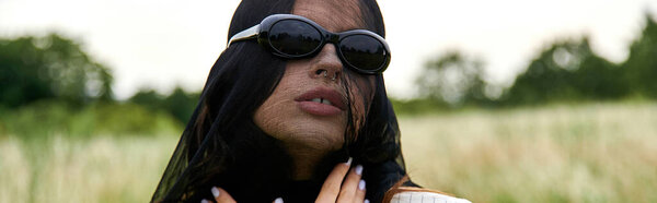 A mysterious woman in a black veil and sunglasses exudes elegance and sophistication in a natural setting.