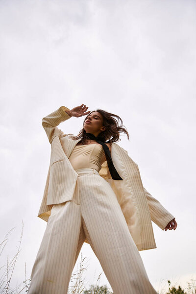 Молодая женщина излучает элегантность в белом костюме и черном галстуке, обнимая летний бриз в безмятежном поле.