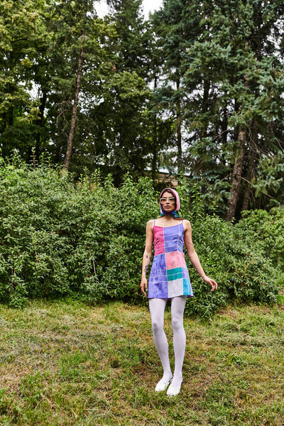 A beautiful young woman in a vibrant dress and sunglasses standing gracefully in the lush green grass, enjoying the summer breeze.