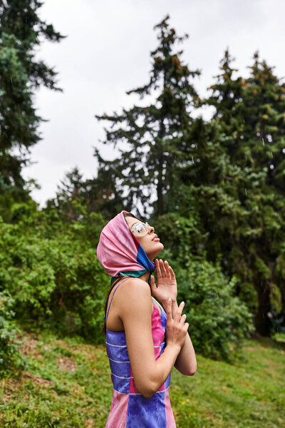 A beautiful young woman in a vibrant blue and pink dress stands gracefully in a field, enjoying the summer breeze.