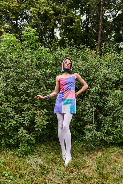 A beautiful young woman in a vibrant dress and sunglasses dances gracefully in a lush green meadow, reveling in the summer breeze.