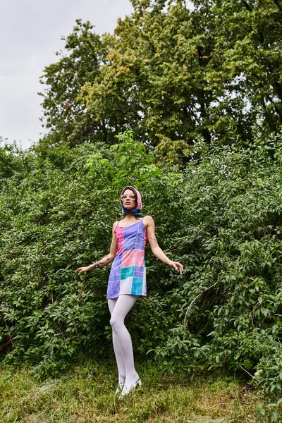 stock image A young woman in a vibrant dress and sunglasses stands in a field with trees in the background, soaking in the summer breeze.