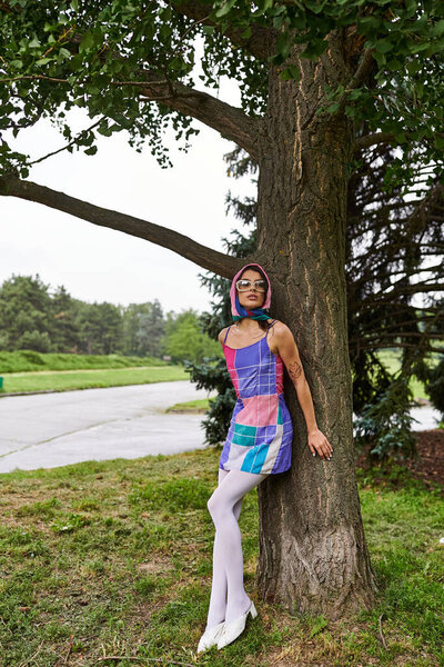 A beautiful young woman in a vibrant dress and sunglasses stands next to a tree, enjoying the summer breeze in a park.