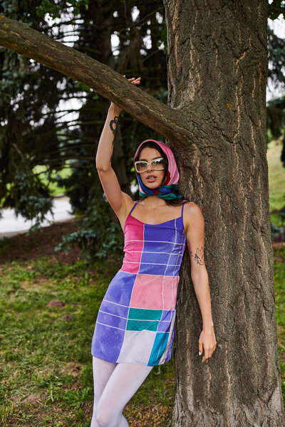 A beautiful young woman in a vibrant dress and sunglasses stands next to a tree, enjoying the summer breeze.