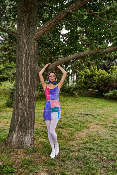A beautiful young woman in a vibrant dress and sunglasses stands next to a tree, enjoying the summer breeze in a tranquil field.
