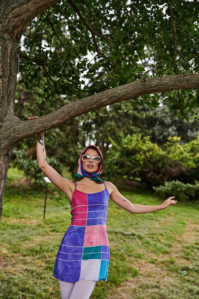 A beautiful young woman in a vibrant dress and sunglasses stands under a tree, basking in the summer breeze.