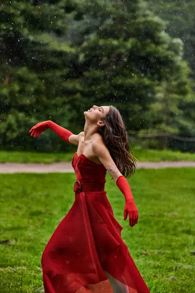 Kırmızı elbiseli güzel genç bir kadın yağmurda zarif bir şekilde duruyor, havaya rağmen zarafet ve duruş sergiliyor..