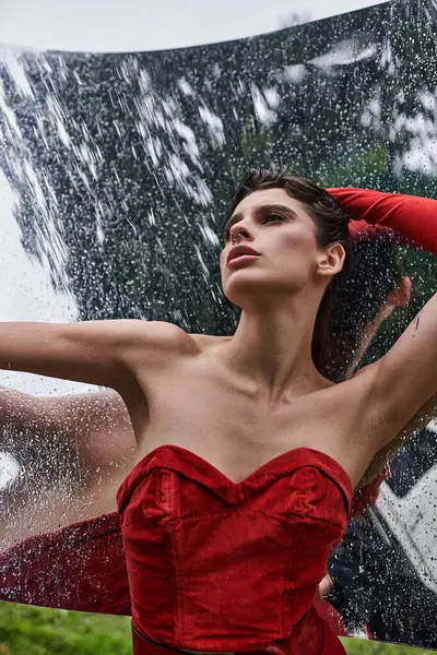 stock image A stunning young woman in a vibrant red dress stands gracefully in the rain, embracing the natural beauty of the moment.