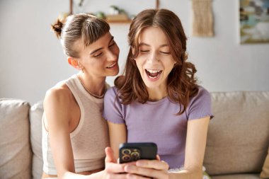 Two women in comfy attire, sitting on a couch, engrossed in a cell phone. clipart