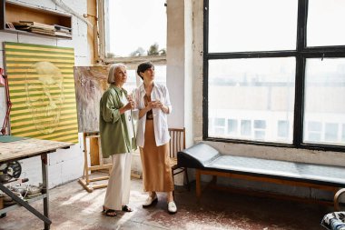 Two mature women sharing a moment of camaraderie in a vibrant art studio. clipart