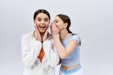 İki güzel, esmer genç kız, spor kıyafetleri içinde, yan yana duruyorlar, elleri yüzlerinde, gri arka planda bir stüdyoda..