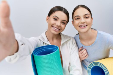 İki güzel ve esmer genç kız, spor giysileri içinde, gri arka planda yoga minderleriyle yan yana duran kız arkadaşları..
