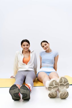 İki güzel ve esmer genç kız, spor kıyafetleri içinde ayaklarını bir mindere koyup stüdyo ortamında otururlar..