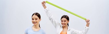 Esmer saçlı iki güzel kadın, gri bir stüdyo arkaplanına karşı spor kıyafetleriyle yan yana duruyorlar..