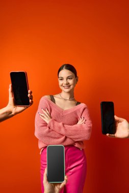 Güzel, esmer, genç bir kız canlı kırmızı bir duvarın önünde duruyor cep telefonlarının yanında, modern bir bağlantı ve teknoloji sergiliyor..