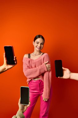 A brunette woman stands before a red wall, near two cell phones in hands of people clipart