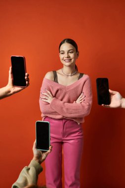 Esmer bir kadın cep telefonlarının yanındaki kırmızı duvarın önünde duruyor, günümüzün dijital çağındaki iletişim ve teknolojisini simgeliyor..