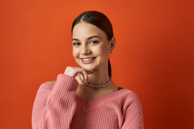 Pembe kazaklı esmer bir kız stüdyo ortamında turuncu arka planda poz veriyor..