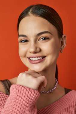 Esmer bir genç kız gülümsüyor, pembe bir kazak giyiyor, turuncu arka planda bir stüdyo ortamında neşe ve mutluluk yayıyor..