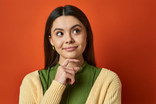 A brunette woman in casual attire looks thoughtful with her hand on her chin, standing against an orange background.