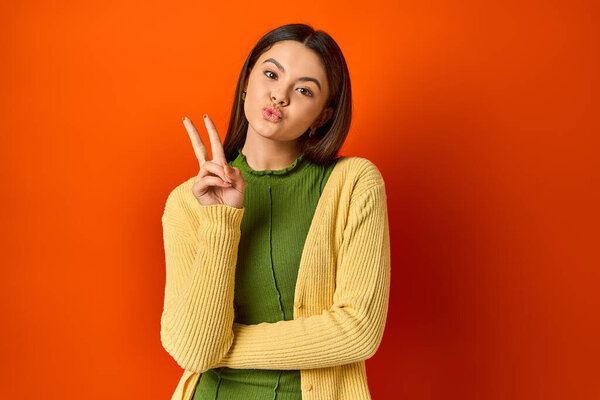 Симпатичная брюнетка-подросток делает знак мира пальцами в студии на оранжевом фоне.