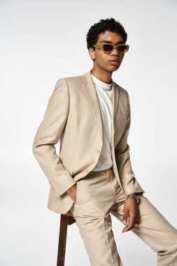 Handsome African American man in stylish sunglasses, sitting on stool in tan suit. clipart