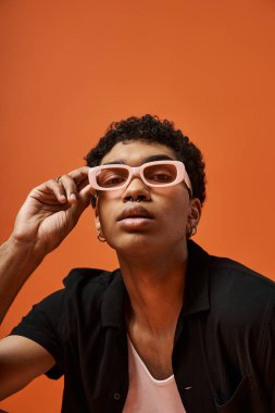 Genç Afro-Amerikan bir adam şık gözlüklü canlı turuncu bir arka plana karşı poz veriyor..