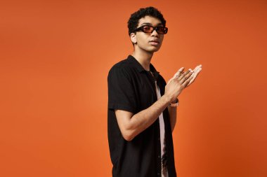 Güneş gözlüklü havalı Afrikalı Amerikalı adam canlı turuncu arka planda rahatça duruyor..