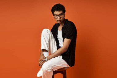 Güneş gözlüklü yakışıklı genç adam parlak turuncu arka planda taburede oturuyor..