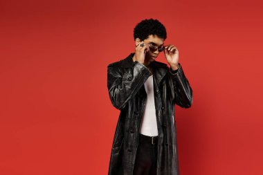 Yakışıklı Afro-Amerikan bir adam şık siyah trençkotuyla çarpıcı bir kırmızı zemine karşı poz veriyor..