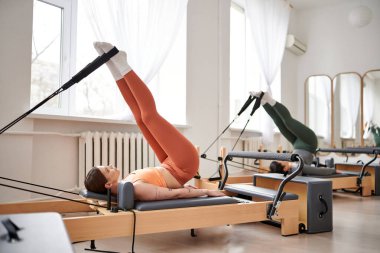 Turuncu bluzlu spor bir kadın pilates dersi sırasında sırtını gerer..
